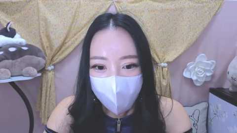 Media: Video of an Asian woman with long black hair, wearing a white surgical mask, dark blue sleeveless top, and sitting against a yellow curtain with stuffed animal decor.