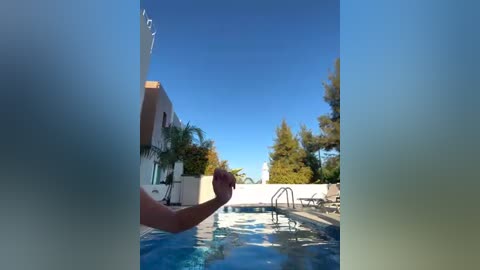 Media: Video of a poolside scene with a person in a red swimsuit holding a ball, surrounded by palm trees, a white building, and clear blue sky.