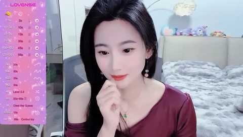 Media: A video of an East Asian woman with long black hair, fair skin, and a slim physique, wearing a burgundy off-shoulder top, posing in a bedroom with a fluffy grey blanket, plush toys, and a pink background.