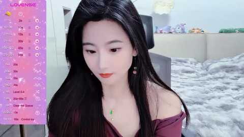 Media: A video of a young East Asian woman with long black hair, wearing a maroon off-shoulder top and earrings, sitting on a bed with a gray blanket, in a softly lit room.