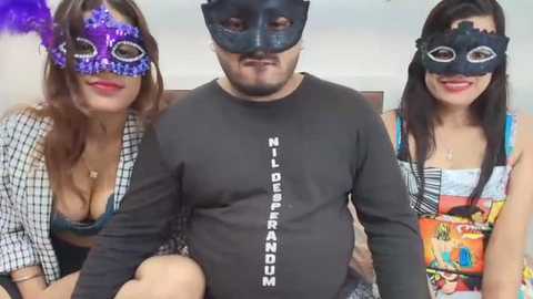 Media: Video of a man with a mustache wearing a black mask, flanked by two women in colorful masks and revealing tops, seated indoors.