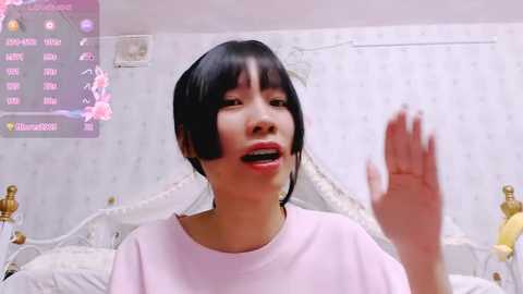 Media: Video of a young Asian woman with short black hair, wearing a white T-shirt, smiling, making a hand gesture. Background features a bed, floral wallpaper, and digital display.