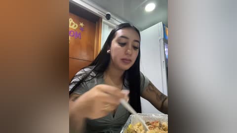 Media: Video of a young woman with long black hair, tattoos, and a gray shirt, eating noodles and fried chicken in a dimly lit room.