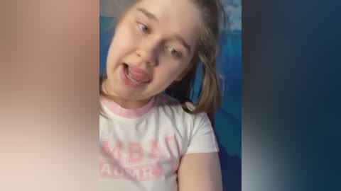 Media: Video of a young Caucasian girl with light skin, blonde hair, and green eyes, wearing a white t-shirt with pink text. She has a playful expression, sticking out her tongue. Background is a gradient of blue and pink.