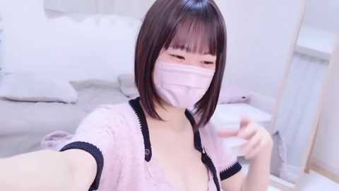 Media: A video of an Asian woman with straight, shoulder-length brown hair and a light pink face mask, wearing a sheer, pink lace robe with black trim, seated on a white couch in a minimalist, brightly lit room.