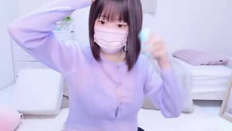 Media: Video of an East Asian woman with straight, shoulder-length black hair, wearing a light purple sweater and a white surgical mask, adjusting her hair in a modern, white-walled bedroom with minimalist decor.