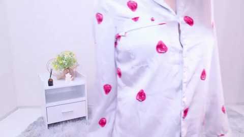 Media: A video of a person wearing a white satin shirt adorned with large, glossy red heart-shaped buttons, standing next to a white nightstand with a small vase of green leaves and a brown bottle.