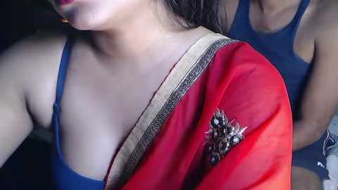 Media: Video of a woman in a red sari with gold embroidery, wearing a blue sleeveless top, with another person partially visible in the background.