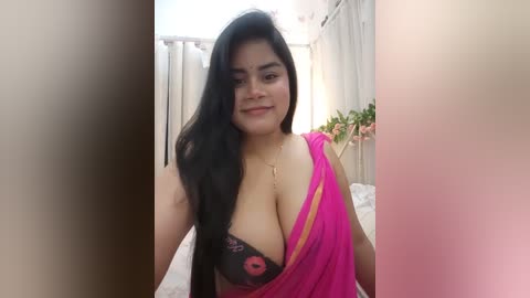 Media: A video of a smiling South Asian woman with long black hair, wearing a pink sari with a black bra peeking out, standing in a brightly lit room with white curtains and floral decor.