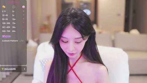 Media: Video of an Asian woman with long black hair, wearing a red halter top, sitting on a white couch in a modern living room.