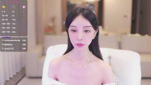 Media: A video of an East Asian woman with long black hair and fair skin, wearing a pearl necklace, seated on a white couch in a modern, softly lit living room.