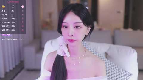 Media: Video of a young Asian woman with long black hair, wearing a white off-shoulder dress, sitting on a couch in a modern living room.