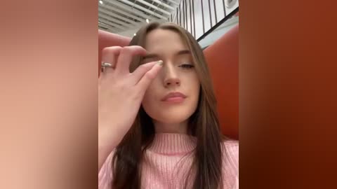 Media: Video of a young woman with long brown hair, wearing a pink sweater, standing in a modern, orange-painted room with black railings. She's covering her eyes with her right hand.