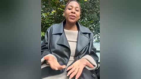 Media: Video of a Black woman with a medium complexion, wearing a black leather jacket over a beige sweater, outdoors with lush greenery in the background. She has a relaxed expression, hands gesturing slightly.