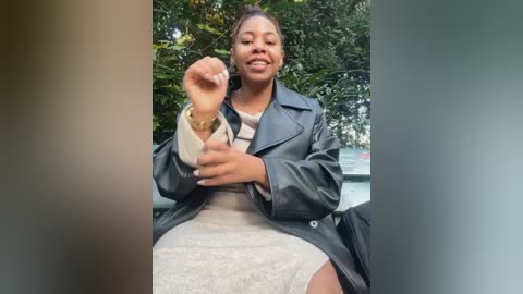 Media: A video of a smiling Black woman with braided hair in a black leather jacket and beige lace dress, gesturing with her right hand outdoors among greenery.