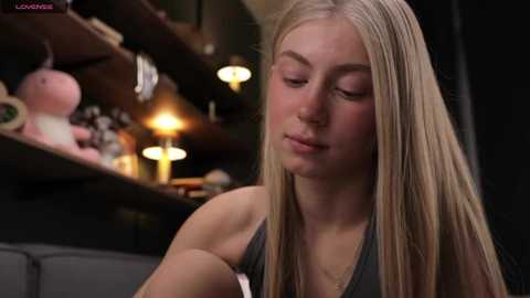 Media: Video of a young blonde woman with straight hair, wearing a gray tank top, looking down at a phone, surrounded by soft lighting and stuffed toys in the background.