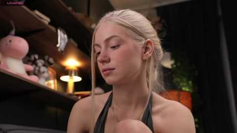 Media: Video of a blonde woman with fair skin and a slender build, wearing a gray tank top, sitting indoors. Background includes shelves with stuffed toys, a lamp, and dark curtains.