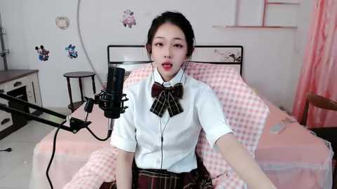 Media: A video of an Asian woman with dark hair, fair skin, and red lipstick, wearing a white blouse and plaid skirt, singing into a microphone in a pastel bedroom.