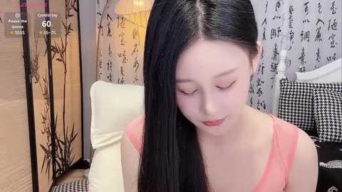 Media: A video of an Asian woman with long black hair, wearing a pink top, sitting on a white couch in a room with black-and-white patterned wallpaper and bamboo screen.