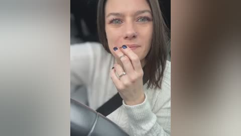 Media: Video of a fair-skinned woman with straight brown hair, light makeup, and blue nail polish, resting her hand on her chin in a car, wearing a white sweater.