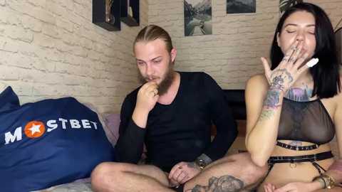 Media: Video of a tattooed man and woman, both topless, sitting on a bed with a blue \"MostBet\" pillow. The woman is covering her mouth with her hand.