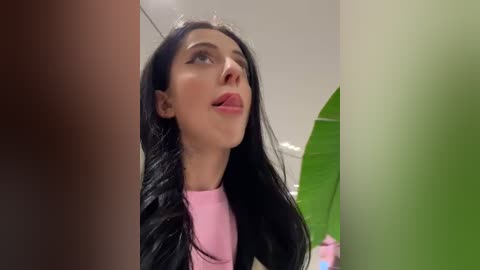 Media: Video of a woman with long black hair, fair skin, wearing pink top, gazing upwards, blurred background with green leaf and pink bottle.