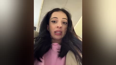 Media: Video of a young woman with long black hair, wearing a pink turtleneck and beige jacket, captured in a blurred, close-up selfie.