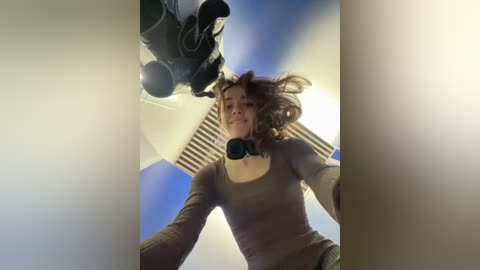 Media: Video of a young woman with curly hair, wearing a brown long-sleeve top, looking up towards a camera mounted on a ceiling, against a bright blue sky.