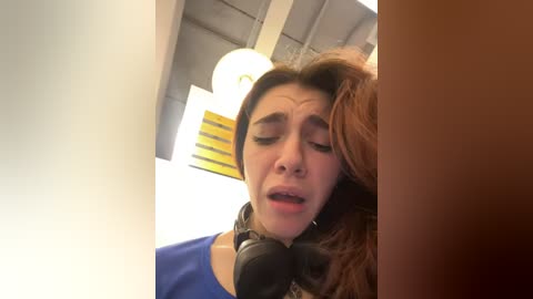 Media: Video of a young woman with long brown hair, wearing a blue shirt, sitting with headphones on, eyes closed, in a dimly lit room with a ceiling fan.