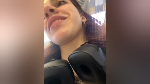 Media: A close-up video of a young woman with light skin, brown hair, and large black headphones. She has a playful expression with her tongue out, captured from a low angle.