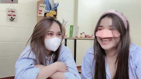 Media: Video of two young East Asian women with light skin, wearing face masks and light blue striped shirts, smiling and looking at the camera, in a simple, brightly lit room.