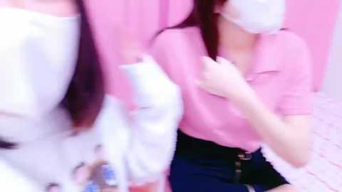 Media: A blurred video shows two women with long hair, wearing pink tops and masks, holding a white object. One has a white dress, the other a black skirt. The background is a pink wall.