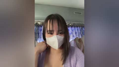 Media: A video of a woman with medium skin tone and straight dark hair, wearing a white face mask, purple top, and blue curtains in the background.