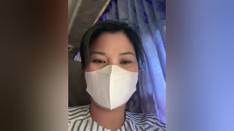 Media: A video of an Asian woman with dark hair, wearing a white face mask and a striped shirt, standing indoors with blue curtains in the background.