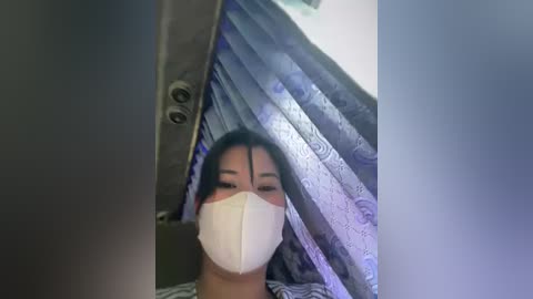 Media: Video of an Asian woman with dark hair, wearing a white mask, standing in a bathroom with a tiled shower curtain and metal hooks.