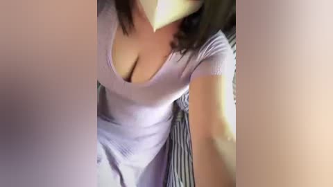 Media: Video of a woman with medium skin tone wearing a low-cut, light purple, ribbed dress, exposing ample cleavage. She has medium-length, dark hair. Background shows striped fabric, likely a bedspread.