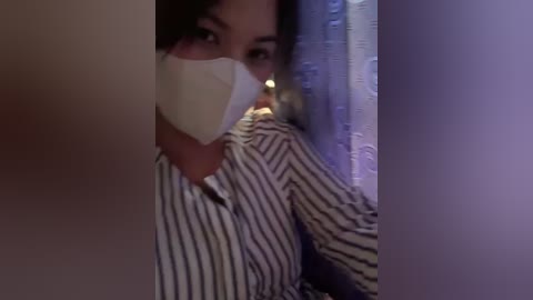 Media: Video of a person in a striped shirt, wearing a white face mask, looking through a doorway. Background features dimly lit curtains and a faint, warm light source.