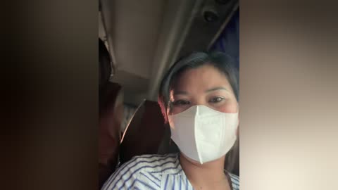 Media: Video of an Asian woman with long black hair, wearing a white mask and a striped shirt, sitting on a plane seat, surrounded by beige curtains and a dark window.