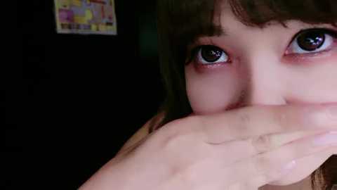 Media: A close-up video of a young Asian girl with big, dark eyes, fair skin, and dark hair, partially covering her mouth with her right hand. The background is dark, with a colorful, abstract painting in the upper left corner.