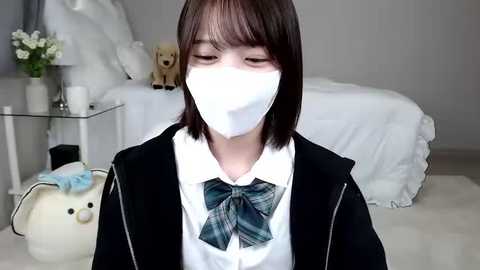 Media: Video of an East Asian girl in a white mask, black jacket, and green plaid bowtie, sitting on a white carpet in a minimalist bedroom with a bed, teddy bear, and flowers.