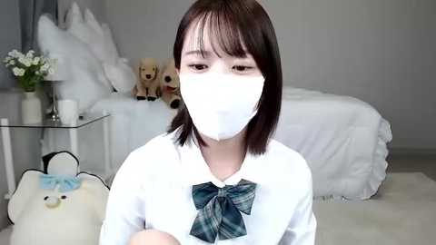 Media: Video of an East Asian woman with short brown hair, wearing a white mask, white blouse, and a green plaid bow, in a minimalist bedroom with a white bed, stuffed toys, and a white pumpkin.