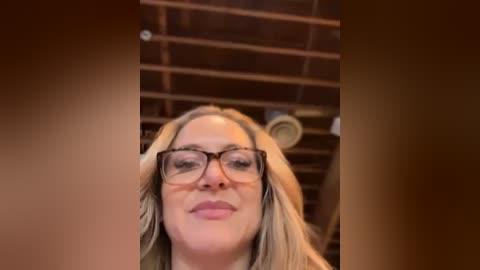 Media: A video of a middle-aged woman with long blonde hair, wearing glasses and a light-colored top, smiling, set against a wooden ceiling background.