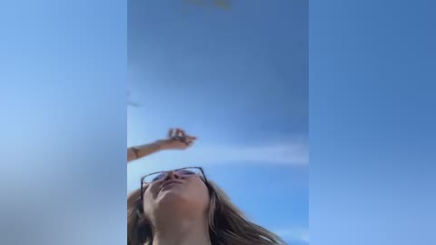 Media: A video of a young woman with long brown hair, wearing glasses, looking up at the clear blue sky, with a slight smile.