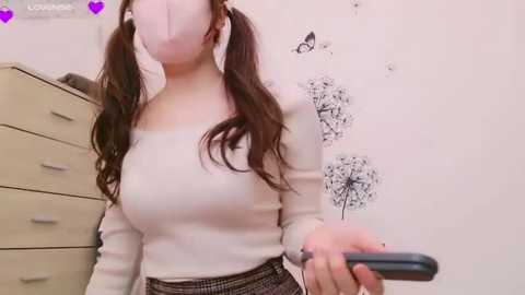 Media: Video of a woman with long brown pigtails wearing a white mask, white sweater, and plaid skirt, holding a smartphone, in a pastel-colored room with dandelion and butterfly wall art.