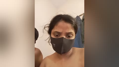 Media: Video of a woman with dark skin, wearing a black mask, taken from the side. She has dark hair and is topless. The background is blurred, featuring a blue curtain and white walls.