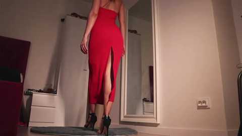 Media: Video of a slender, light-skinned woman with long hair, wearing a red, strapless, high-cut gown with a thigh slit, black high heels, and standing in a modern, dimly lit room with white walls and a mirror.