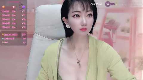 Media: Video of an East Asian woman with short black hair, wearing a green top and a necklace, seated in a white chair. Background shows a pink wall and a blurred figure.