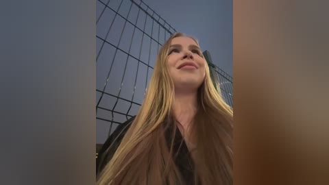 Media: Video of a blonde woman with straight hair, smiling, standing in front of a wire fence against a blue sky.