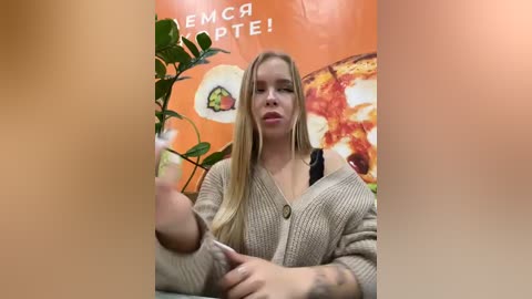 Media: Video of a blonde woman in a beige knit cardigan, holding a pizza slice, against a vibrant orange wall with pizza posters and green plants.