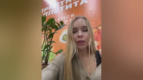 Media: Video of a blonde woman with long hair, fair skin, and wearing a beige cardigan, standing in front of an orange wall with Russian text and a plant.